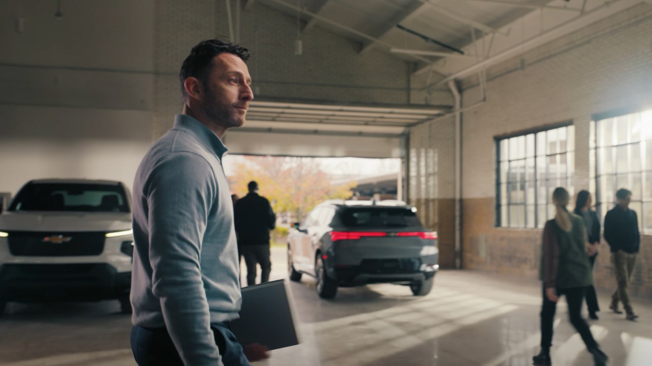 A thumbnail for the OnStar EV Reimbursement Video displaying a man walking past two GM vehicles