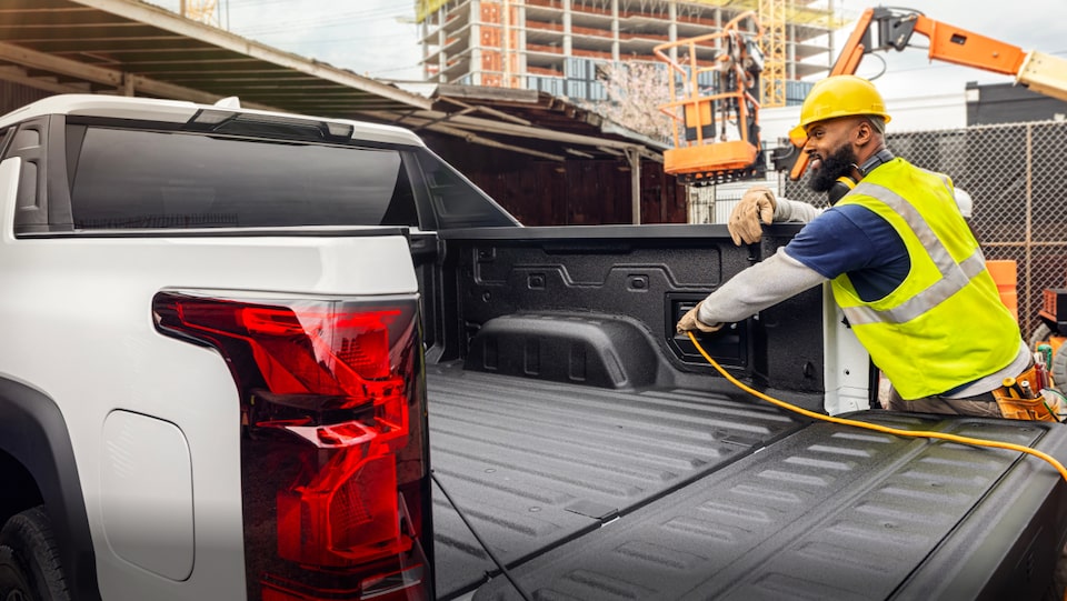 An Exterior view of a Construction Worker connecting a charger cable within the trunk area of the  Silverado EV at a Construction Site.