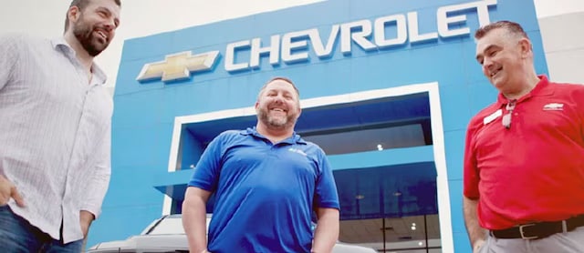 Three men standing in front of a Chevrolet dealership