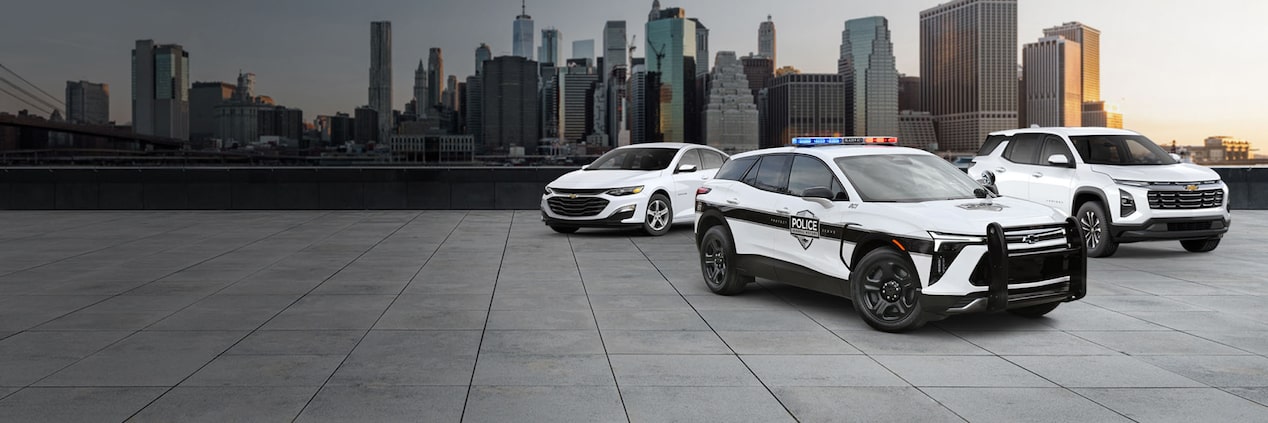 A group of  white chevrolet vehicles with a police vehicle centered within the image on a natural skyline building background.